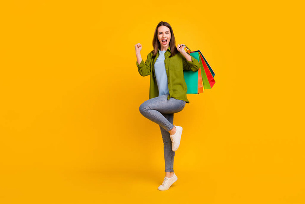Photo of excited lucky young woman dressed green shirt holding bargains rising fist smiling isolated yellow color background. - Foto, imagen