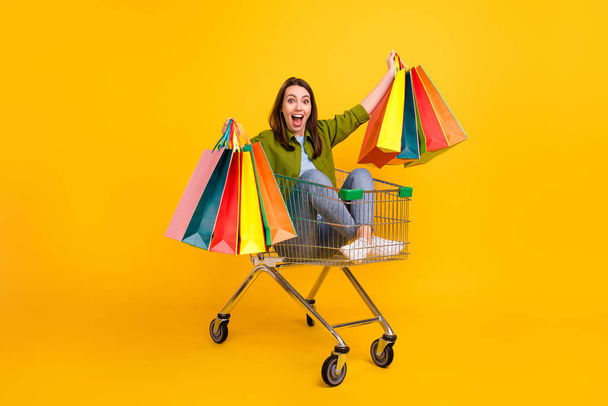 Photo of funny excited young woman dressed green shirt holding bargains riding cart smiling isolated yellow color background. - Foto, immagini