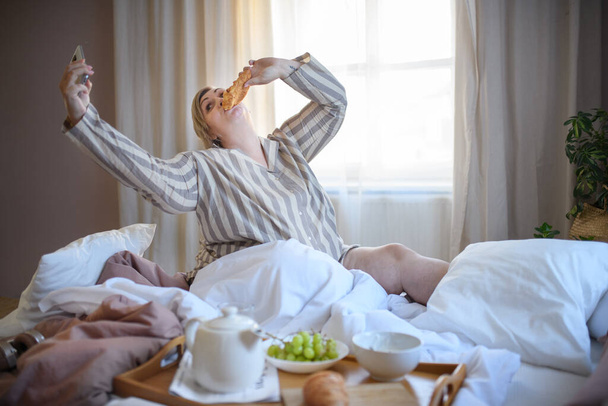 A happy overweight woman with headphones and smartphone having breakfast in bed at home. - Fotografie, Obrázek