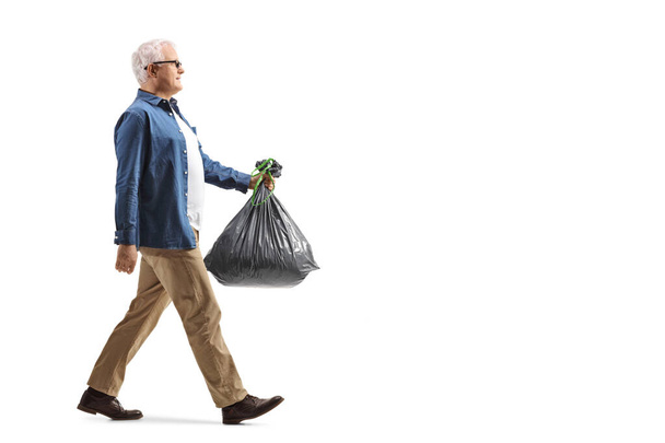 Full length profile shot of a mature man walking and carrying a plastic waste bag isolated on white background - Foto, immagini
