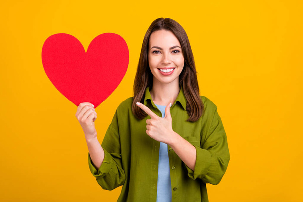 Photo of sweet charming young woman dressed green shirt smiling pointing finger big red heart isolated yellow color background. - Fotografie, Obrázek