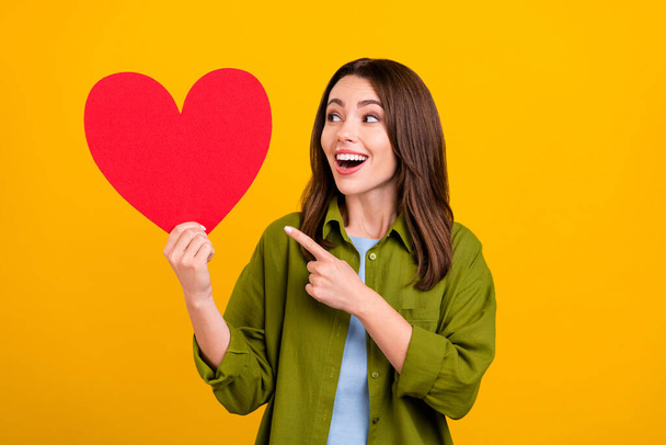 Photo of charming excited woman dressed green shirt smiling looking pointing finger big red heart isolated yellow color background. - Zdjęcie, obraz