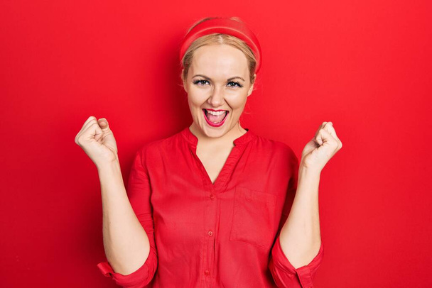 Young blonde woman wearing casual red shirt screaming proud, celebrating victory and success very excited with raised arms  - Fotoğraf, Görsel