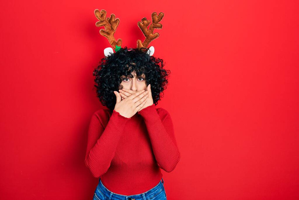 Молодая женщина с Ближнего Востока, одетая в милые рождественские рога северного оленя, потрясена, закрывая рот руками за ошибку. секретное понятие.  - Фото, изображение