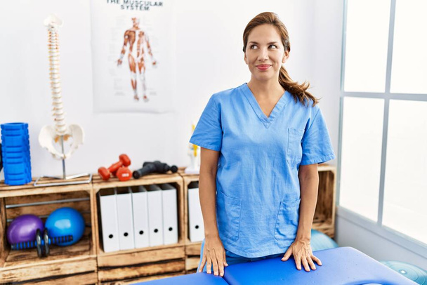 Middle age hispanic physiotherapist woman working at pain recovery clinic smiling looking to the side and staring away thinking.  - Foto, Imagem