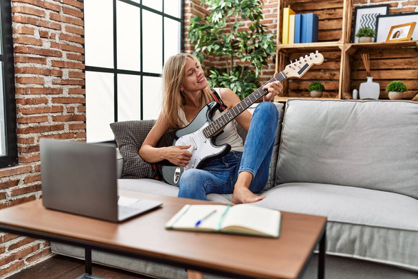 jong blond vrouw glimlachen zelfverzekerd spelen elektrische gitaar thuis - Foto, afbeelding
