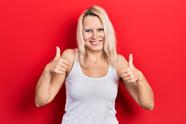 Belle femme blonde caucasienne portant un signe de succès de t-shirt blanc décontracté faisant un geste positif avec la main, le pouce levé souriant et heureux. expression joyeuse et geste gagnant.  - Photo, image