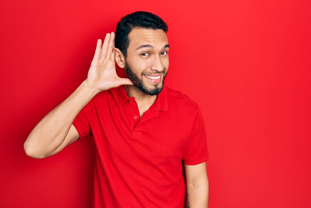 Spanyol férfi szakállal, alkalmi piros pólóban, aki fültől fülig mosolyog, hallgatva a pletykákat vagy pletykákat. süketség fogalma.  - Fotó, kép