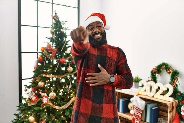 Afrikkalainen amerikkalainen joulupukin hattu päässä, seisoo joulukuusen vieressä nauraen sinulle, osoittaen sormella kameraa käsin, häpeän ilme  - Valokuva, kuva