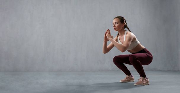A sports woman in fashion sport clothes squatting doing sit-ups in gym, over gray background - Foto, afbeelding