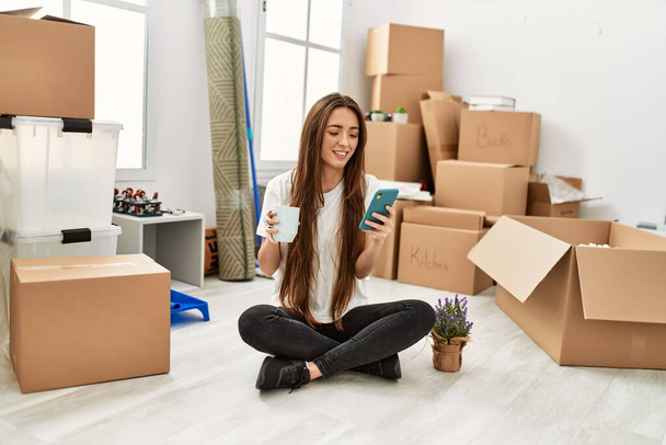Young hispanic woman using smartphone drinking coffee sitting on floor at new home - Photo, Image