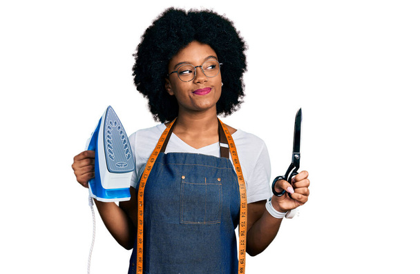 Junge afrikanisch-amerikanische Schneiderin Designerin mit Atelierschürze, Eisen und Schere in der Hand, lächelnd zur Seite schauend und nachdenkend wegstarrend.  - Foto, Bild