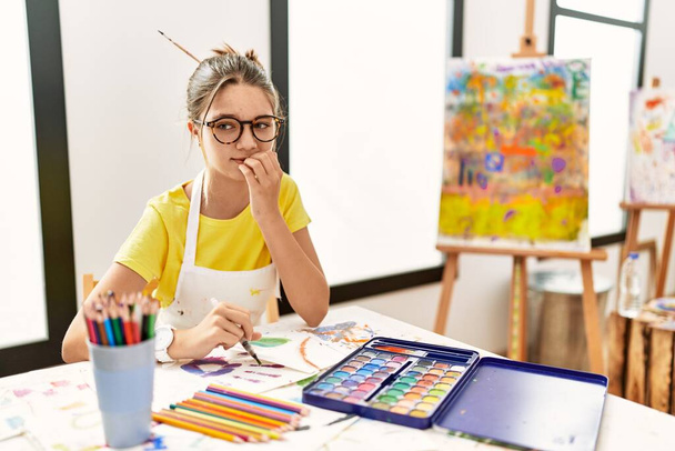 Jeune adolescente brune au studio d'art à l'air stressée et nerveuse avec les mains sur les ongles mordants. problème d'anxiété.  - Photo, image