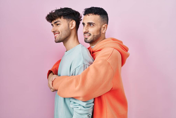 Jeune couple gay hispanique debout sur fond rose regardant vers le côté, pose de profil relax avec visage naturel et sourire confiant.  - Photo, image