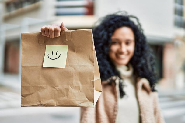 Nuori latino nainen hymyilee luottavainen tilalla ottaa pois paperipussi kadulla - Valokuva, kuva