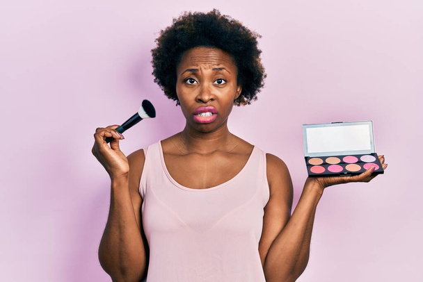 Νεαρή Αφροαμερικανή γυναίκα που κρατάει πινέλο μακιγιάζ και κοκκινίζει ανίδεη και μπερδεμένη έκφραση. έννοια της αμφιβολίας.  - Φωτογραφία, εικόνα