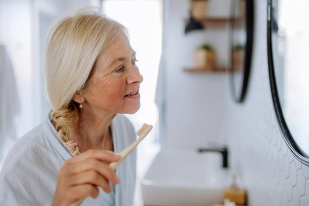 A b eautiful senior woman in bathrobe brushing teeth with eco wooden toothbrush inbathroom, sustainable lifestyle. - Фото, изображение