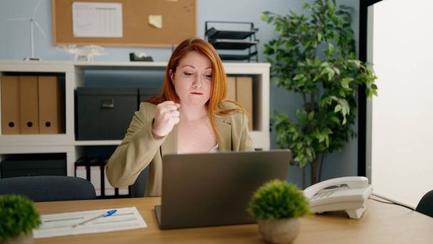 Молодая рыжая деловая женщина злится, используя ноутбук, работающий в офисе - Фото, изображение