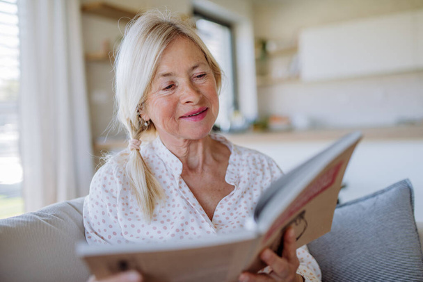 Una donna anziana seduta sul divano e che legge un libro a casa - Foto, immagini