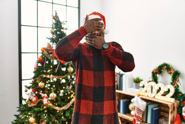 Αφροαμερικάνος που φορούσε καπέλο Σάντα Κλάους, στεκόταν δίπλα στο χριστουγεννιάτικο δέντρο καλύπτοντας τα μάτια και το στόμα με τα χέρια, έκπληκτος και σοκαρισμένος. απόκρυψη συγκίνησης  - Φωτογραφία, εικόνα