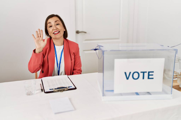 Красива жінка середнього віку на політичних виборах, сидячи на виборах, відмовляючись від голосування, каже привіт щасливий і усміхнений, дружній вітальний жест
  - Фото, зображення