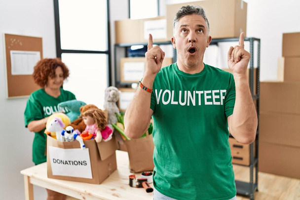 Hombre de mediana edad vistiendo camiseta voluntaria en las donaciones de pie sorprendido y sorprendido mirando hacia arriba y señalando con los dedos y los brazos levantados.  - Foto, imagen