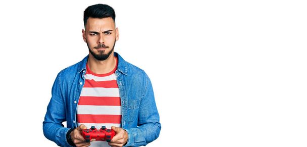 Молодой латиноамериканец с бородой играет в видеоигры держа контроллер скептически и нервно, хмурясь расстроен из-за проблемы. отрицательный человек.  - Фото, изображение