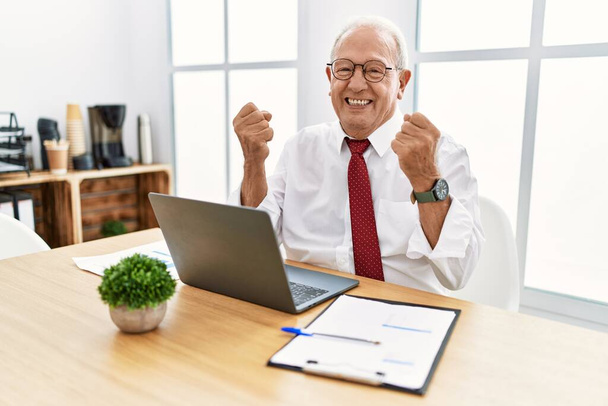 Ein älterer Mann, der im Büro mit einem Computer-Laptop arbeitet, ist sehr glücklich und aufgeregt und macht Siegergeste mit erhobenen Armen, lächelt und schreit nach Erfolg. Festkonzept.  - Foto, Bild