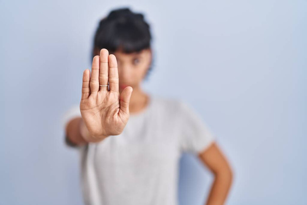 Jeune femme hispanique portant t-shirt décontracté sur fond bleu faire arrêter de chanter avec la paume de la main. expression d'avertissement avec geste négatif et sérieux sur le visage.  - Photo, image