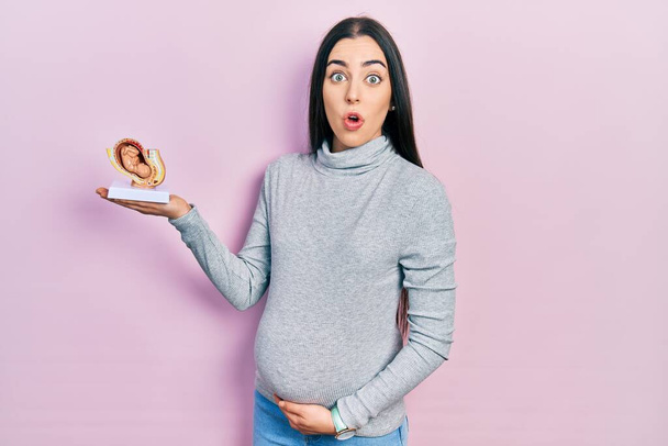 Beautiful woman with blue eyes expecting a baby, holding anatomic fetus scared and amazed with open mouth for surprise, disbelief face  - Zdjęcie, obraz