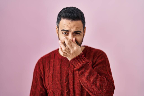 Молодой латиноамериканец с бородой в обычном свитере на розовом фоне чует что-то вонючее и отвратительное, невыносимый запах, затаив дыхание с пальцами на носу. плохой запах  - Фото, изображение