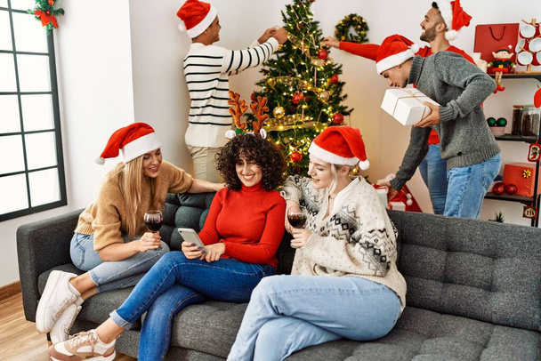クリスマスを祝う若者のグループ。3人の女性がワインを飲み、自宅でスマートフォンを使う. - 写真・画像