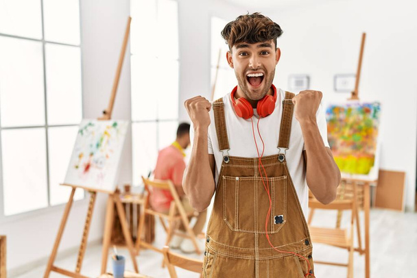 Sanat stüdyosundaki genç İspanyol adam şaşkınlık ve şaşkınlık içinde kollarını kaldırıp gözleri açık bir şekilde başarıyı kutluyor. Kazanan konsept.  - Fotoğraf, Görsel