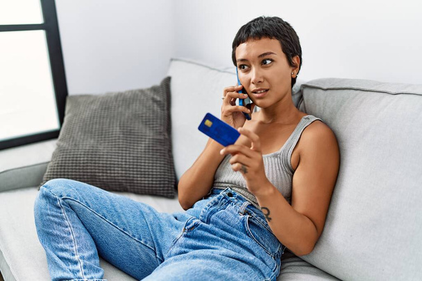 Jeune femme hispanique parlant sur le smartphone tenant la carte de crédit à la maison - Photo, image