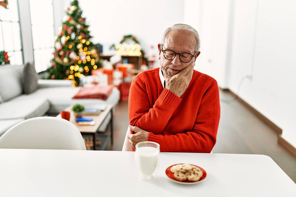Idős férfi ősz hajjal ül az asztalon a cookie-kat karácsonyfa gondolkodás fáradtnak tűnik, és unatkozik a depresszió problémák keresztbe karok.  - Fotó, kép