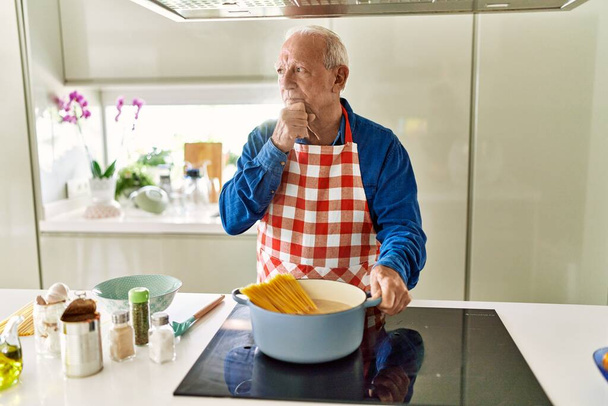 Homem sênior com cabelos grisalhos cozinhar espaguete em casa cozinha rosto sério pensando em questão com a mão no queixo, pensativo sobre ideia confusa  - Foto, Imagem