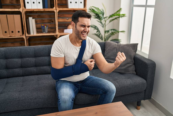 Jeune homme hispanique avec la barbe portant le bras sur fronde assis à la thérapie consulter ennuyé et frustré criant de colère, criant fou de colère et la main levée  - Photo, image