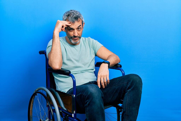 Handsome middle age man with grey hair sitting on wheelchair worried and stressed about a problem with hand on forehead, nervous and anxious for crisis  - Photo, Image
