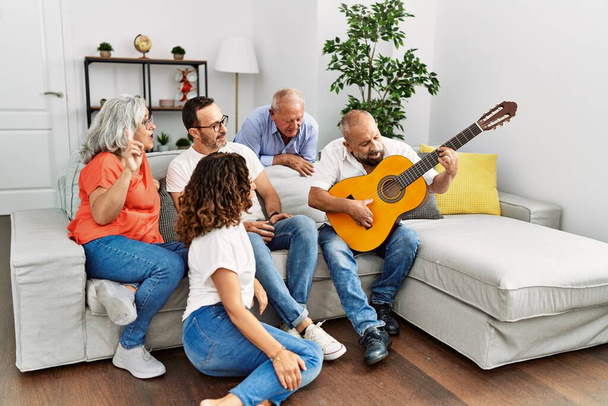 Grupo de amigos de mediana edad que tienen fiesta tocando la guitarra clásica sentados en el sofá en casa. - Foto, imagen