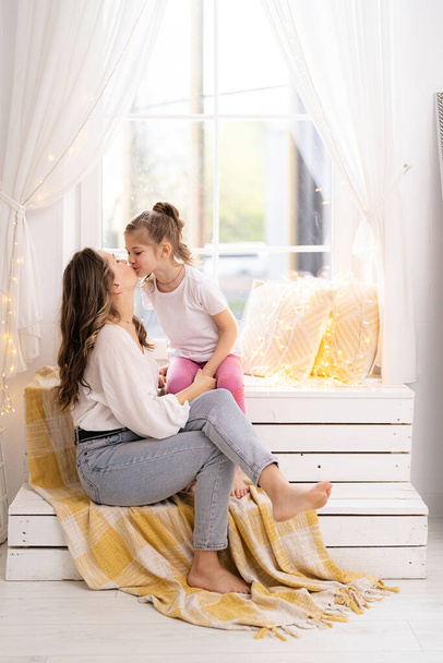 Unity mother and daughter concept. Girl hugging her mother, sitting on the window, showing her love at home - Photo, Image