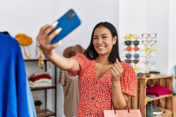 Νεαρή Λατίνα γυναίκα κάνει selfie από το smartphone κρατώντας ρούχα στο κατάστημα ρούχων - Φωτογραφία, εικόνα