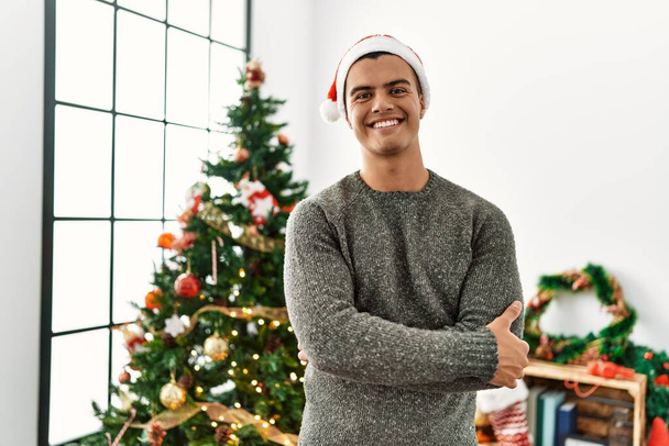 Young hispanic man smiling confident with arms crossed gesture standing by christmas tree at home - Fotografie, Obrázek