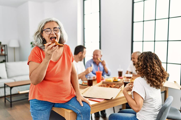 Orta yaşlı bir grup mutlu bir şekilde gülümseyen İtalyan pizzasını evde masada oturuyor. - Fotoğraf, Görsel