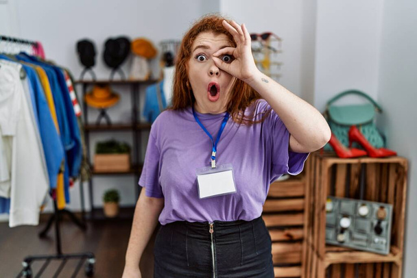 Young redhead woman working as manager at retail boutique doing ok gesture shocked with surprised face, eye looking through fingers. unbelieving expression.  - Foto, afbeelding