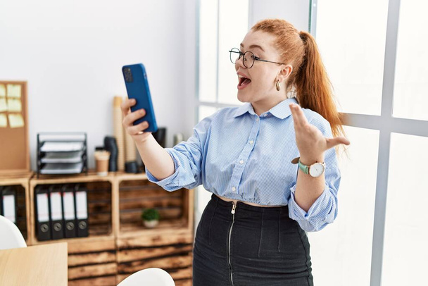 Young redhead woman smiling confident using smartphone at office - Фото, изображение