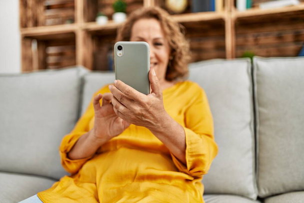 Középkorú kaukázusi nő usin okostelefon ül a kanapén otthon. - Fotó, kép