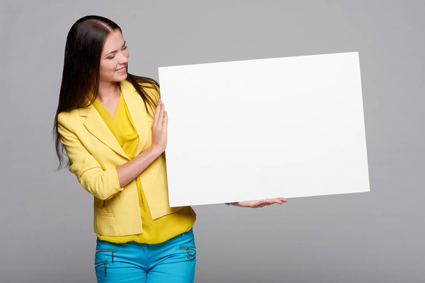 Happy smiling girl in yellow and blue, holding empty whiteboard, looking at blank copy space at board - Photo, image