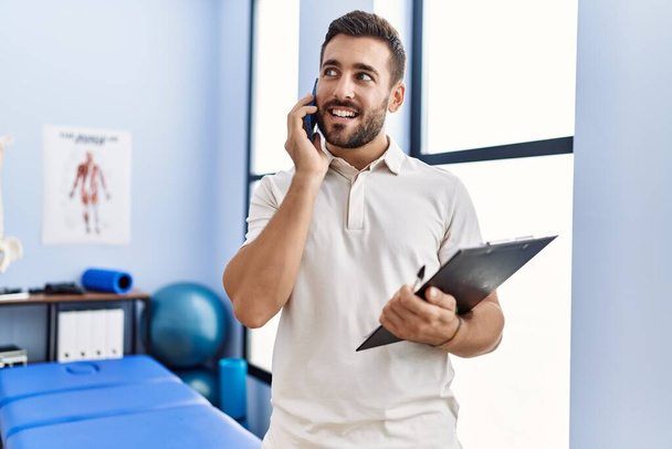 Joven hombre hispano con uniforme de fisioterapeuta hablando en el smartphone de la clínica - Foto, Imagen