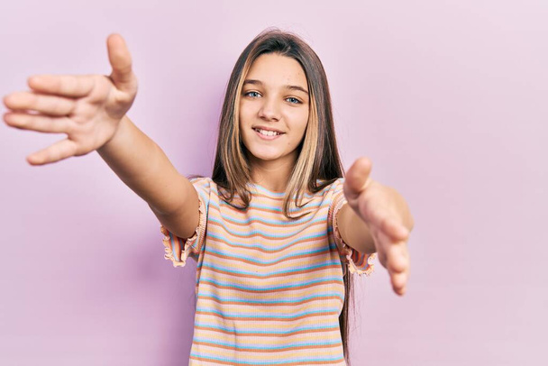Молодая брюнетка в повседневной полосатой футболке смотрит на камеру, улыбаясь с распростертыми объятиями для объятий. веселое выражение, охватывающее счастье.  - Фото, изображение