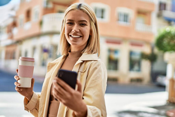 Młoda blondynka za pomocą smartfona pije kawę w mieście. - Zdjęcie, obraz